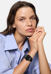 Schwarze rechteckige Smartwatch mit digitalem Display am Handgelenk, getragen über einem hellblauen Hemd mit Kragen. Glatter Gummiriemen, minimalistisches Design.