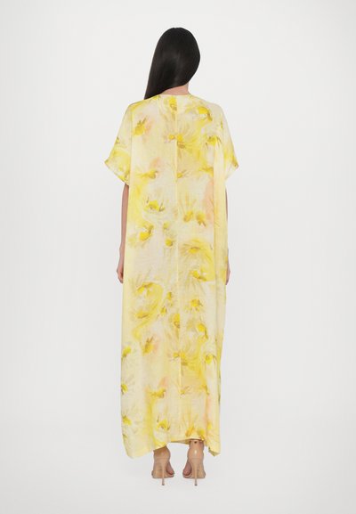 Lång gul kaftan med blommönster, korta ärmar och lös passform, tillverkad av lättviktig tyg. Sömmen är något rundad på sidorna.