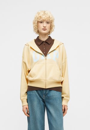 Persoon met krullend blond haar draagt een beige GAP hoodie met ritssluiting over een bruin shirt en een blauwe spijkerbroek, staand tegen een effen achtergrond.