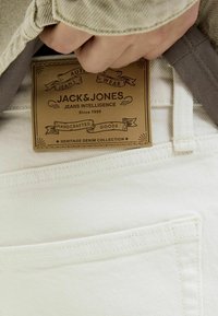 Jeans in denim bianco con etichetta in pelle che mostra "JACK & JONES" e dettagli decorativi. Zoom sui punti e sul design delle tasche.