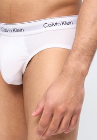 Άνδρας που φοράει λευκά εσώρουχα Calvin Klein με ορατό λογότυπο στη ζώνη, το χέρι ακουμπάει στο μηρό, πάνω σε ανοιχτό γκρι φόντο.