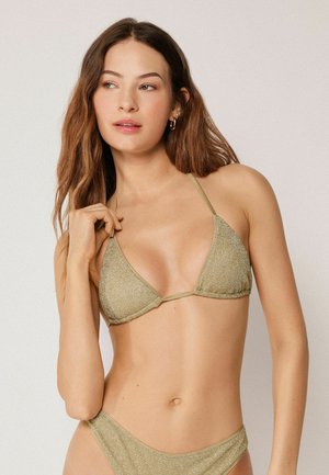 Ung kvinde med langt brunt hår iført en guldfarvet glitrende trekants-bikinitop og bund, poserede mod en ensfarvet lys baggrund.