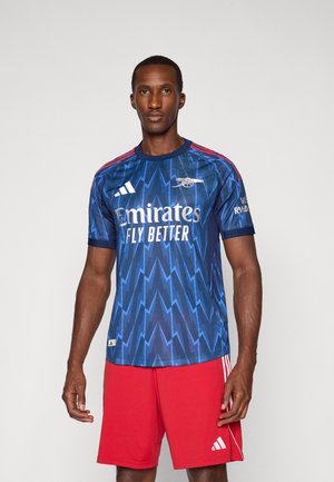 Blaues Fußballtrikot mit Blitzmuster, roten Akzenten an den Schultern, weißem "Emirates"-Logo, getragen mit roten Shorts mit drei Streifen.