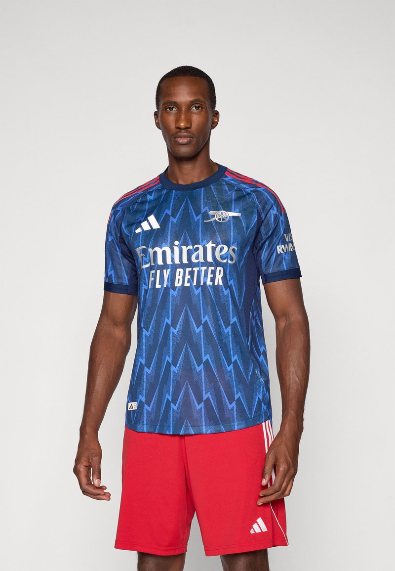 Blå fodboldtrøje med lynmønster, røde detaljer på skuldrene, hvid "Emirates" logo, båret med røde shorts med tre striber.