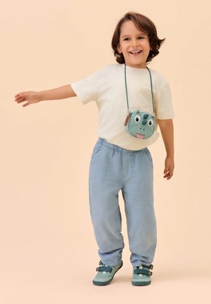 Lächendes Kind, das ein weißes T-Shirt, hellblaue Hose, türkisfarbene Schuhe und eine Umhängetasche in Dinosaurierform trägt und mit ausgestrecktem Arm steht.