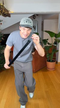 Ceinture en cuir noir tenue par une personne portant un t-shirt gris et un pantalon gris. En arrière-plan, un canapé marron et des plantes en pot.