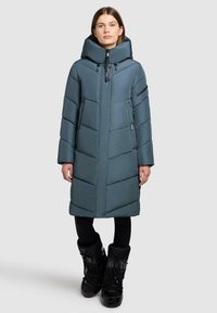 Kvinde iført en lang teal farvet dunjakke med høj krave og sorte vinterstøvler, stående mod en ensfarvet lys baggrund.