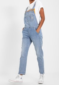 Pechera de mezclilla azul claro con tirantes ajustables, bolsillos frontales y un diseño de pierna ajustada; combinada con una camiseta gráfica blanca y zapatillas blancas.