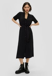 Czarna sukienka midi z guzikami, z paskiem w talii, krótkimi rękawami i głębokim dekoltem w kształcie litery V. Teksturalna tkanina z płynącą sylwetką. Ubrane czarne buty wojskowe.