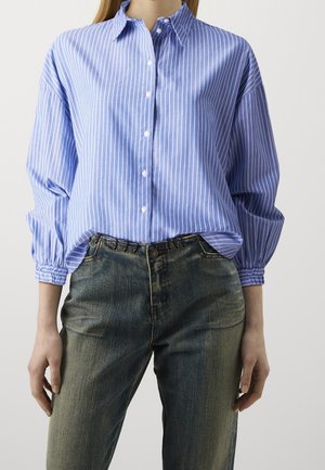 Femme portant une chemise boutonnée à rayures verticales bleues et blanches avec des manches bouffantes et un jean vert foncé délavé.