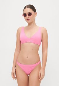 Rožinė banguota bikinio kostiumėlis su giliu V formos dekolte viršumi ir atitinkamais apatiniais. Modelis vilki rožinius akinukus prieš paprastą foną.