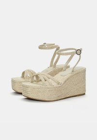 Sandales compensées beiges avec une tige en corde tressée et des brides au niveau de la cheville, présentant une semelle en jute texturé et un accent métallique sur la boucle de la bride.