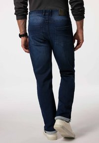 Jeans de un azul oscuro con un diseño ajustado, que presentan piernas rectas, dos bolsillos traseros y un parche de cuero en la cintura. Con puños doblados y zapatos blancos.