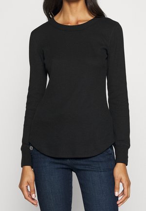 Femme portant un haut noir près du corps à manches longues avec un col rond et un jean bleu foncé sur un fond uni.