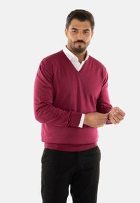 Maglione a V in un profondo bordeaux, realizzato in materiale morbido, con polsini e orlo a coste. Indossato sopra una camicia bianca con colletto.