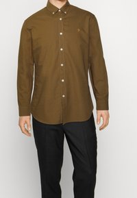 Camicia marrone con bottoni frontali e colletto appuntito, con bottoni argentati e un piccolo logo ricamato sul petto, abbinata a pantaloni neri.