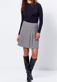 Haut noir à manches longues avec détail en nœud, associé à une jupe plissée grise à carreaux. Bottes en cuir noir jusqu'aux genoux. Textures lisses, coupe ajustée.