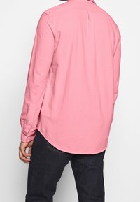 Osoba v růžové košili s dlouhým rukávem a tmavých džínách, zobrazená zezadu na jednobarevném bílém pozadí.