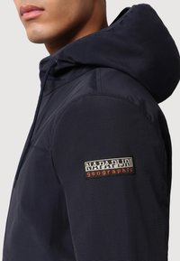 Giacca a vento blu navy con cuciture visibili e superficie testurizzata. Presenta un'etichetta con il marchio sulla spalla sinistra in colori a contrasto.