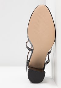 Sandale à talon haut en cuir verni noir vue de la semelle avec un dessous beige lisse et un talon carré, sur fond blanc.