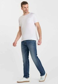 T-shirt en coton blanc avec une poche à l'avant, associé à un jean bleu. La tenue est complétée par des baskets blanches avec des accents beiges.
