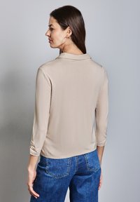 Camicia beige aderente con colletto, maniche a tre quarti e dettagli arricciati ai polsini. Abbinata a jeans blu a gamba larga.