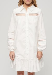 Abito camicia bianco in cotone, con chiusura a bottoni, colletto a punta, maniche lunghe a sbuffo con dettagli in pizzo e orlo arricciato.