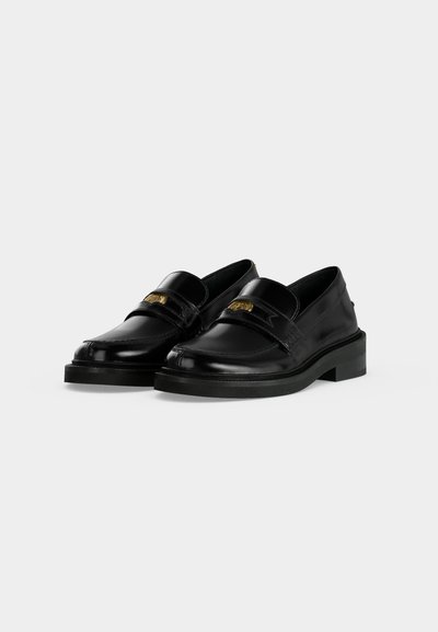 Paire de mocassins en cuir noir avec un emblème décoratif doré sur le dessus, avec une semelle légèrement surélevée et un bout arrondi.
