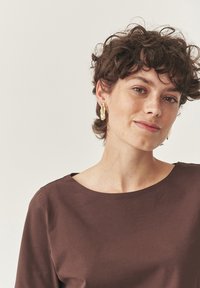 Haut marron à manches courtes avec un large col. Le sujet porte des boucles d'oreilles en or. Tissu texturé. Design simple et minimaliste. Fond clair.