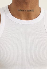Top alb cu dungi, fără mâneci, cu decolteu rotund, având un tatuaj vizibil pe gât care spune "crede în tine".
