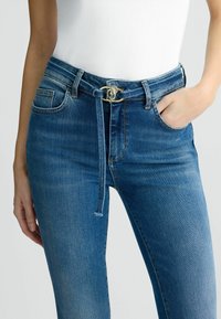 Jeans in denim blu con vita alta, tasche frontali e una cintura con fibbia circolare dorata. Presentano un dettaglio in vita legato e una leggera scoloritura.