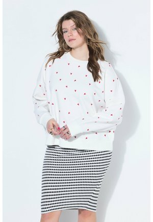 Femme aux cheveux ondulés portant un sweat blanc avec de petits cœurs rouges et une jupe noire et blanche à motifs jusqu'aux genoux, debout contre un mur blanc.