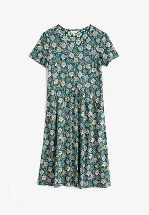 Kvinde iført tealblå blomsterprintet midi-kjole med korte ærmer og samlet talje samt brune åbne flade sandaler, stående mod en hvid væg.