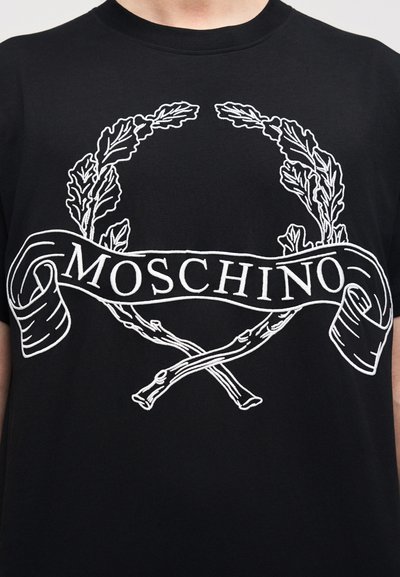 Μαύρο μπλουζάκι με λευκά κεντημένα σχέδια: ένα στεφάνι δάφνης με σταυρωτά ραβδιά και μια κορδέλα που φέρει τη λέξη "MOSCHINO".