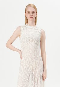 Čipkasta obleka v off-white barvi, ki se ponaša z teksturiranim, nabranim izrezom, brezrokavnim dizajnom in obširnimi vzorčnimi izrezi po celotni tkanini.