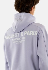 Felpa viola chiaro con cappuccio, con scritta bianca sulla schiena: "PROJECT X PARIS MEMBERS' CLUB" e ulteriori dettagli di design.