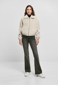 Lodden, kremfarvet lynlåsjakke med krave og elastiske manchetter, parret med grå flagrende jeans og hvide sneakers.