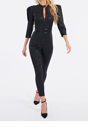 Vrouw die een strakzittend zwart jumpsuit draagt met driekwart mouwen, een ceintuur in de taille, een ritssluiting aan de voorkant en zwarte hoge hakken met enkelbandje.