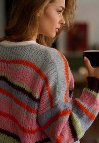 Žena se světle hnědými vlasy, která má na sobě barevný pruhovaný pletený svetr, drží černý hrníček blízko obličeje a dívá se doprava.