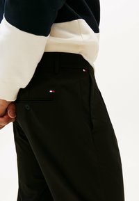 Czarne spodnie o eleganckim wzornictwie, z drobną teksturą i małym haftowanym logo w trzech kolorach na kieszeni. Widoczny detal guzika.