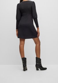 Robe noire à manches bouffantes, tissu texturé, silhouette ajustée évasée, longueur genou. Associée à des bottines noires à talons.