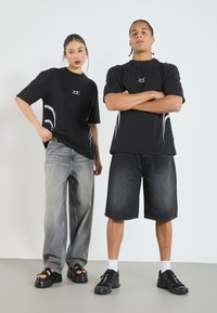 Två personer bär svarta grafiska t-shirts med vita detaljer. Den ena kombinerar en t-shirt med ljusgrå vida byxor, medan den andra har på sig shorts.