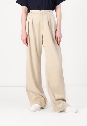 Beige vide benbukser med folder, med en glat bomuldsstruktur og høj talje, parret med hvide sneakers.