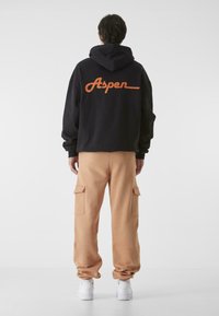 Fekete kapucnis pulóver narancssárga "Aspen" felirattal a hátsó részen, bézs cargo nadrággal, oldalzsebekkel, és fehér sportcipővel párosítva.