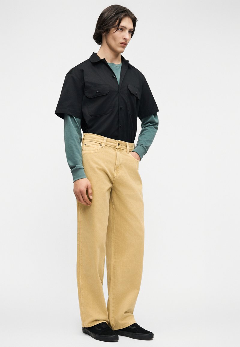 Joven de pie con la mano en el bolsillo, vistiendo pantalones anchos beige, camisa verde de manga larga y camisa negra de manga corta abotonada.