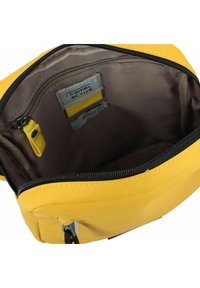 Gelbe Stofftasche mit glatter Textur, Reißverschlussverschluss und braunem Innenfutter. Verfügt über ein Markenschild und eine kleine gelbe Stoffschleife.