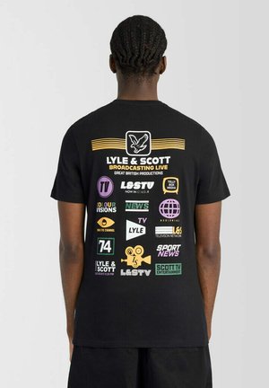Person mit geflochtenem Haar trägt ein schwarzes T-Shirt mit bunten TV- und Medienlogos und Text auf der Rückseite über die Übertragung von Lyle & Scott.