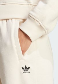 Ljusbeige Adidas sweatshirt med ribbad krage och ärmslut. Noterbara detaljer inkluderar ett sidofack och en liten svart logotyp.