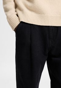 Bež pleteni pulover z rebrastimi manšetami, v kombinaciji s črnimi nabranimi hlačami, ki prikazujejo gladko teksturo in čiste linije v bližnjem pogledu.