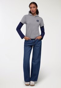 Maglietta a maniche corte grigia con cappuccio e maniche lunghe blu, caratterizzata da una grafica sul petto, abbinata a jeans in denim blu a gamba larga e sneakers bianche.